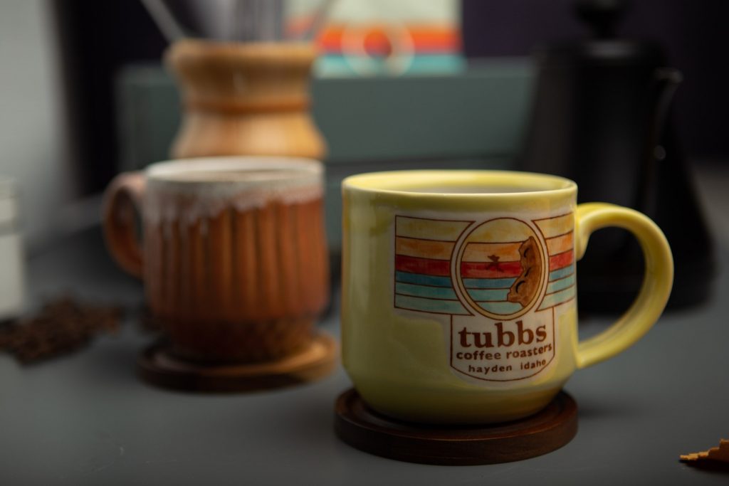 handcrafted, yellow coffee mug featuring the tubbs coffee logo, with additional handmade pottery style mugs in the background